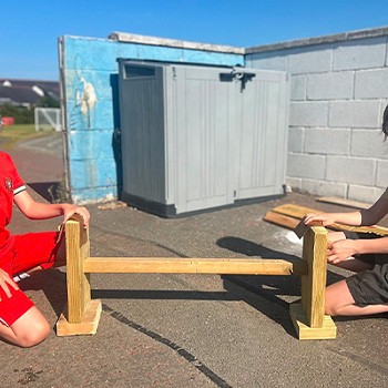 Dau bachgen yn helpu ei gilydd i adeiladu meinciau i fynd gyda'r bwrdd.