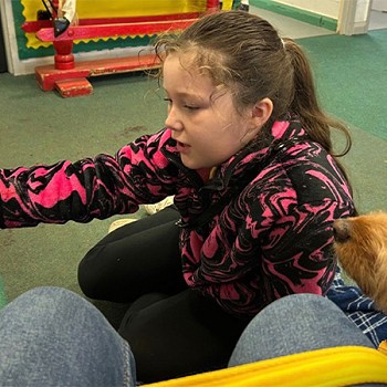 A girl reading a book to Ned while sitting on the floor in the classroom