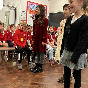 Tair merch yn cymryd rhan yn gwasanaeth blwyddyn 3 a 4 gyda plant yn y cefndir